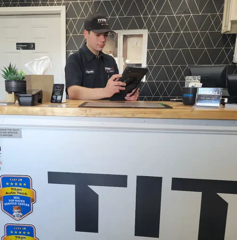 About Titan Auto Tech European & Import Auto Repair. A man in a black uniform and cap stands behind a reception counter, using a tablet. The counter has a plant, tissues, and awards displayed, with TITAN partially visible on the front. The background features a geometric wall pattern.
