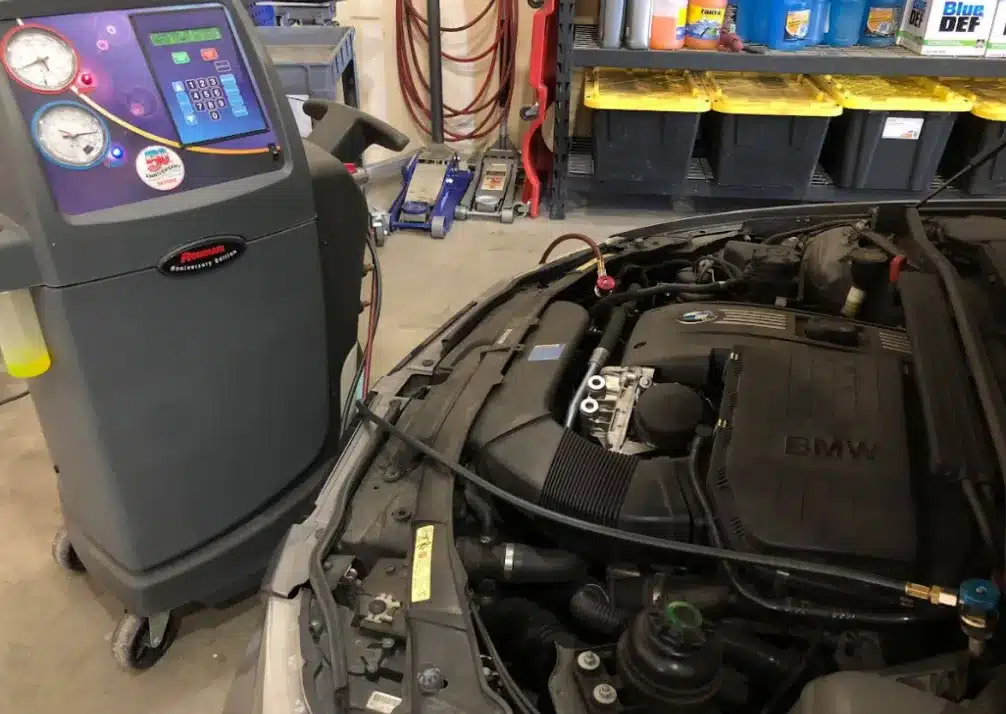 A/C & Heater Repair In Castle Rock CO At Titan Auto Tech European & Import Auto Repair. An automotive air conditioning service machine is connected by hoses to the engine bay of a BMW car in a workshop, with tools and shelves visible in the background.