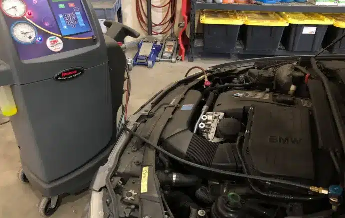 A/C & Heater Repair In Castle Rock CO At Titan Auto Tech European & Import Auto Repair. A machine is connected to the engine of a BMW car in a garage, likely for air conditioning servicing or maintenance. Tools and bins are visible in the background.