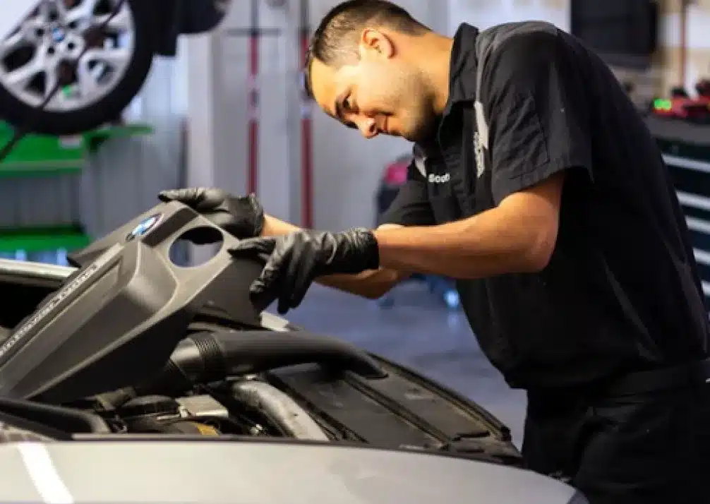 Auto Repair In Castle Rock CO At Titan Auto Tech European & Import Auto Repair. A mechanic wearing black gloves and a black uniform inspects or repairs the engine of a car in an auto repair shop. A vehicle is visible on a lift in the background.