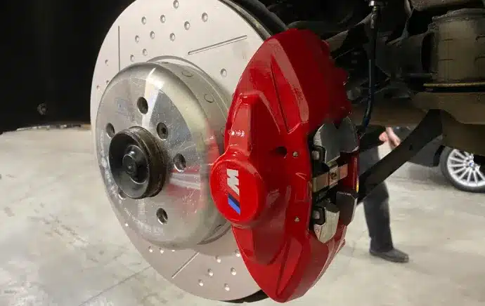 Brake Repair In Castle Rock CO At Titan Auto Tech European & Import Auto Repair. A close-up of a cars front wheel assembly showing a large, silver ventilated brake disc and a red M-branded brake caliper, with part of a person and another car visible in the background.