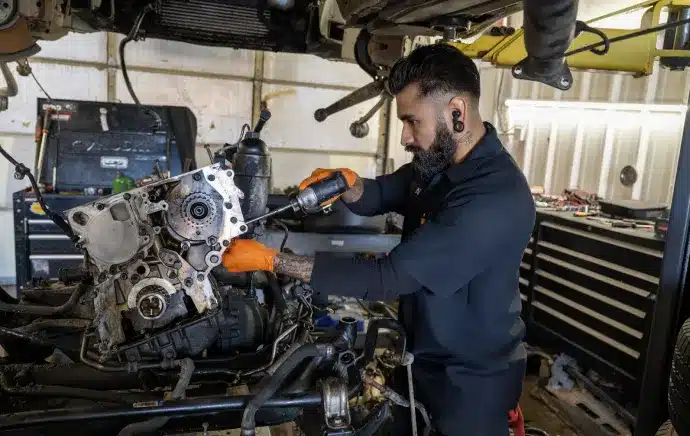 Engine Repair & Replacement In Castle Rock CO At Titan Auto Tech European & Import Auto Repair. A mechanic with tattoos and ear gauges, wearing orange gloves, works with a power tool on an engine in a workshop filled with tools and equipment.
