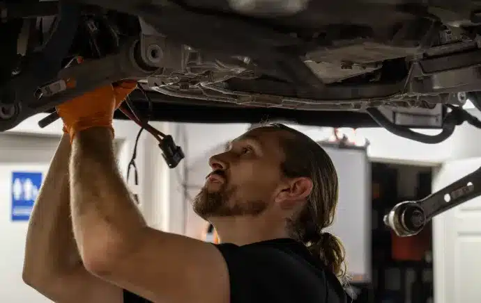 Fleet Maintenance In Castle Rock CO At Titan Auto Tech European & Import Auto Repair. A mechanic with a beard and ponytail, wearing orange gloves, works underneath a raised vehicle in a garage or workshop.