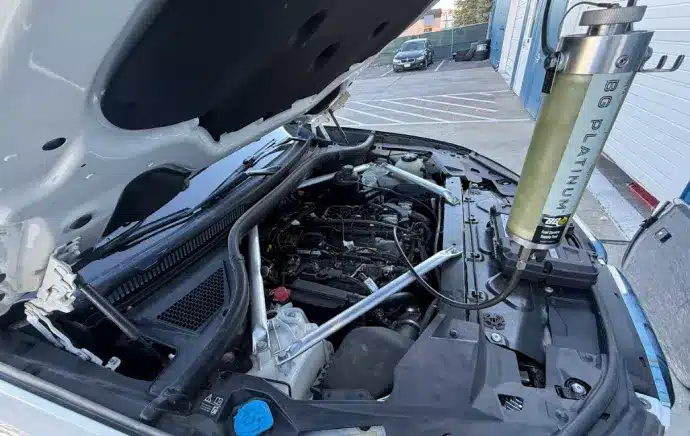 Oil Change In Castle Rock CO At Titan Auto Tech European & Import Auto Repair. A car with its hood open in a parking lot, showing the engine and a BG Platinum tool attached, likely for engine maintenance or cleaning.