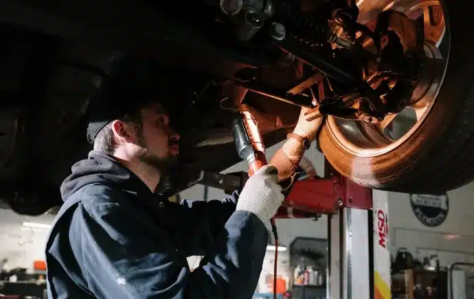 Suspension Repair In Castle Rock CO At Titan Auto Tech European & Import Auto Repair. A mechanic wearing gloves and a cap works under a lifted vehicle, using a power tool to repair the suspension or wheel area in an auto repair shop.
