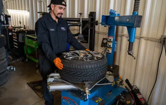 Tire Repair & Services In Castle Rock CO At Titan Auto Tech European & Import Auto Repair. A mechanic wearing orange gloves and a black uniform uses a tire changing machine to fit a tire onto a wheel inside an auto repair shop.