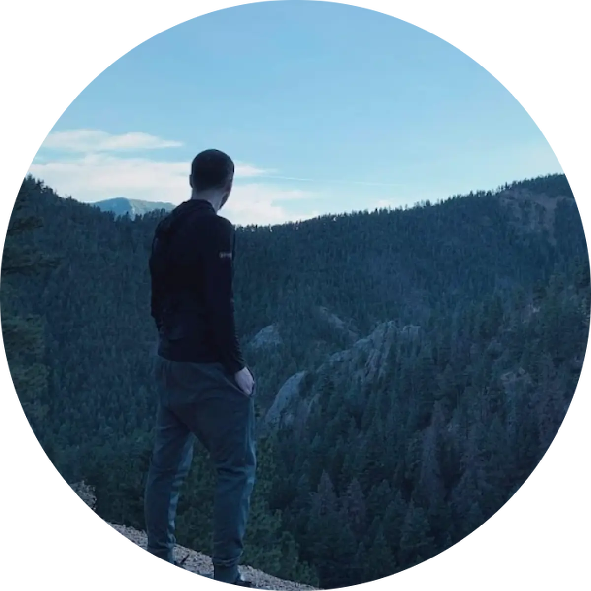 David Service Manager Auto Repair In Castle Rock CO At Titan Auto Tech European & Import Auto Repair. A person in dark clothing stands on a rocky ledge, looking out over a forested mountain landscape under a blue sky. The image is framed in a circular crop.