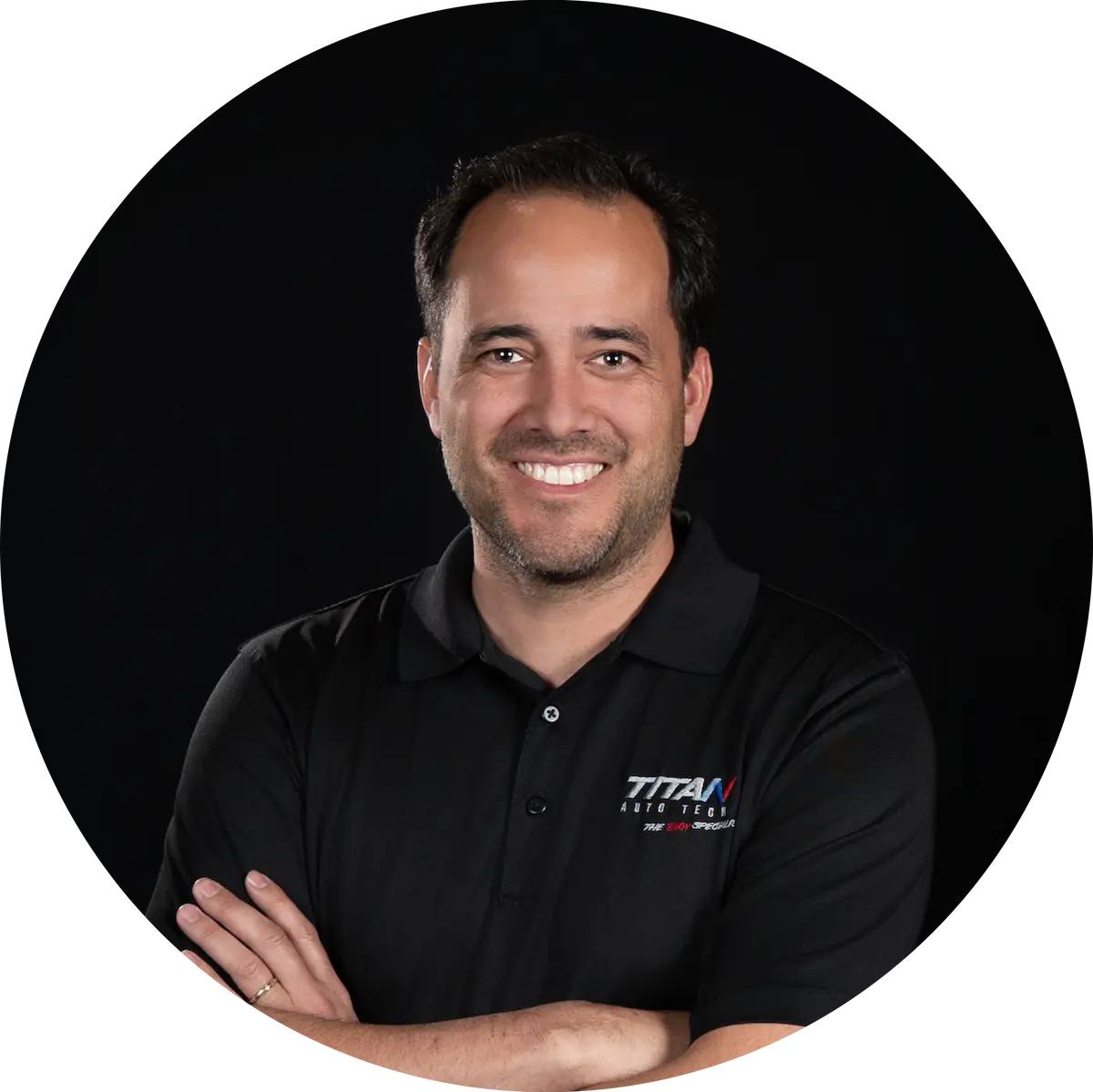 Scott Founder & President Auto Repair In Castle Rock CO At Titan Auto Tech European & Import Auto Repair. Smiling man with dark hair in a black polo shirt with TITAN logo, standing with arms crossed against a plain black background.