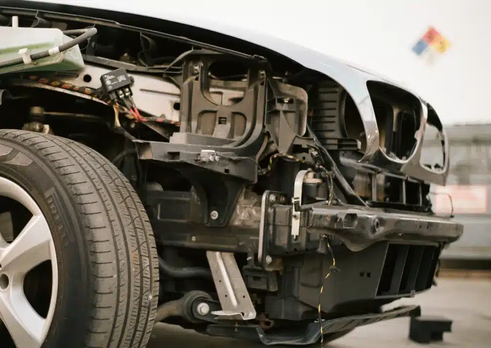 BMW Repair In Castle Rock CO At Titan Auto Tech European & Import Auto Repair. A close-up view of the front left side of a car with its bumper and headlights removed, exposing internal mechanical components and wiring. The car tire and part of the wheel are visible.