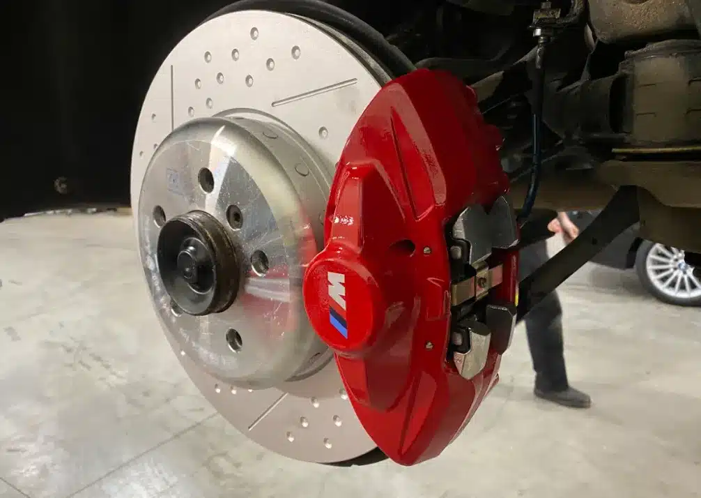Brake Repair In Castle Rock CO At Titan Auto Tech European & Import Auto Repair. A close-up view of a car’s front brake assembly, showing a ventilated and drilled brake rotor with a bright red caliper featuring an M logo, inside a garage setting.