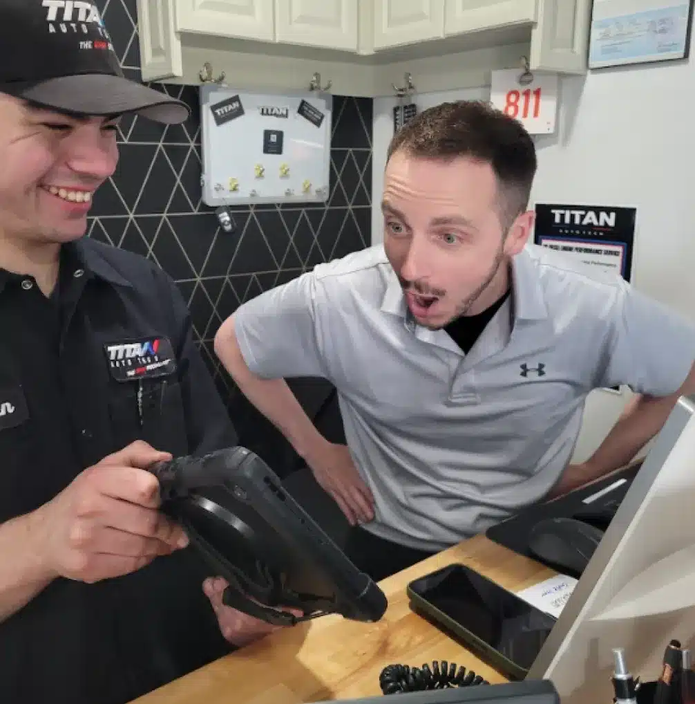 Auto Repair Career In Castle Rock CO At Titan Auto Tech European & Import Auto Repair. Two men at a counter smile and react with surprise while looking at a tablet device. One wears a black uniform and cap, the other a light gray polo shirt. Office items and a computer are visible in the background.