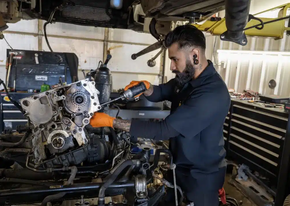 Engine Repair & replacement In Castle Rock CO At Titan Auto Tech European & Import Auto Repair. A mechanic with tattoos and ear gauges, wearing orange gloves, uses a power tool to work on a car engine inside an auto repair shop. Tools and equipment are visible in the background.