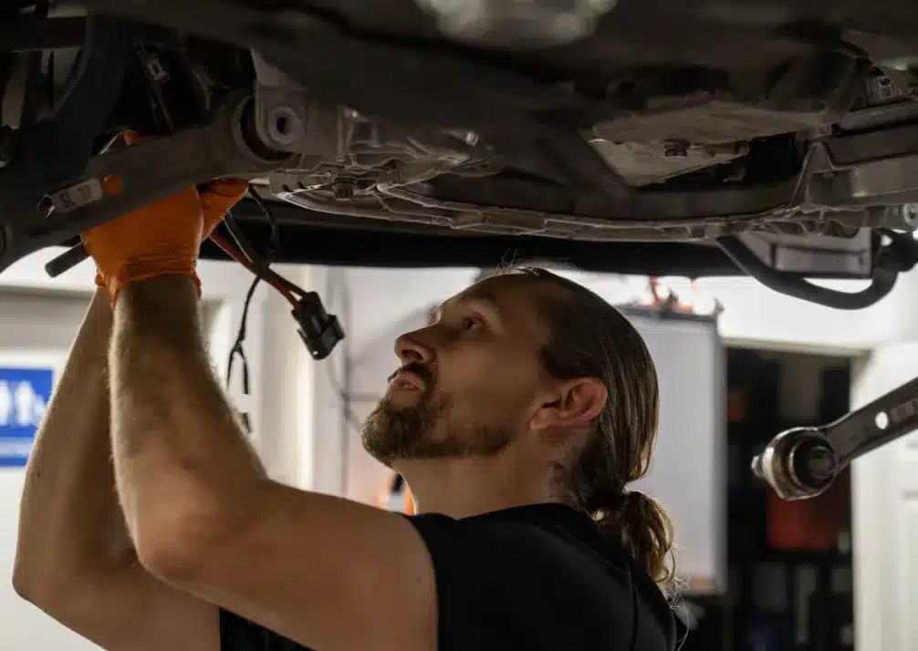 Fleet Maintenance In Castle Rock CO At Titan Auto Tech European & Import Auto Repair. A mechanic with a beard and ponytail, wearing orange gloves, works underneath a vehicle in a garage, focusing intently on repairs.