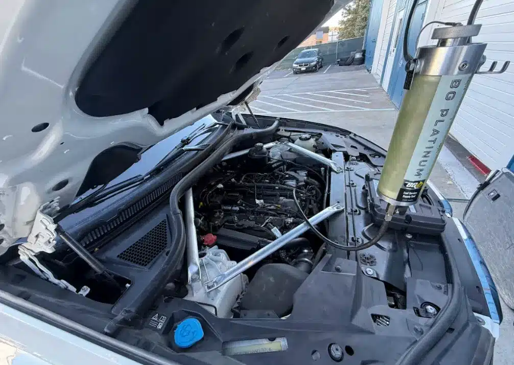 Oil Change In Castle Rock CO At Titan Auto Tech European & Import Auto Repair. Close-up view of a car engine with the hood open, showing a BG Platinum tool connected for maintenance. The vehicle is parked in a lot near a building with a garage door.