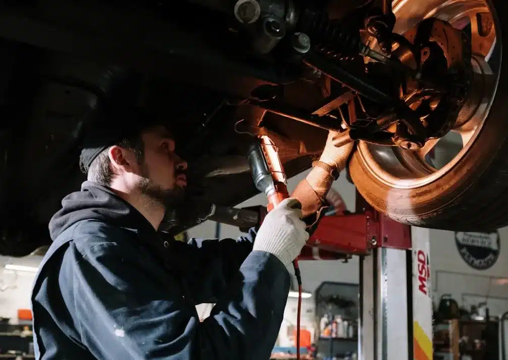 Suspension Repair In Castle Rock CO At Titan Auto Tech European & Import Auto Repair. A mechanic in a dark jacket and gloves works under a raised vehicle, using a flashlight and tool to inspect or repair parts near the wheel in a garage workshop.