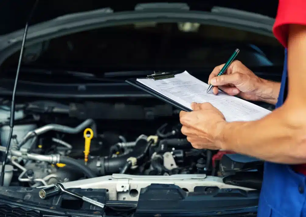 Vehicle Inspections In Castle Rock CO At Titan Auto Tech European & Import Auto Repair. A person holding a clipboard and pen stands in front of an open car hood, inspecting and writing notes, likely performing a vehicle maintenance check or inspection.