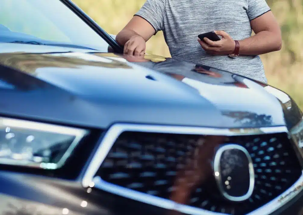 Acura Repair In Castle Rock CO At Titan Auto Tech European & Import Auto Repair. A person in a gray t-shirt leans on the hood of a black car, holding a smartphone in one hand. The image focuses on the cars front grille and headlight, with the person partially visible.