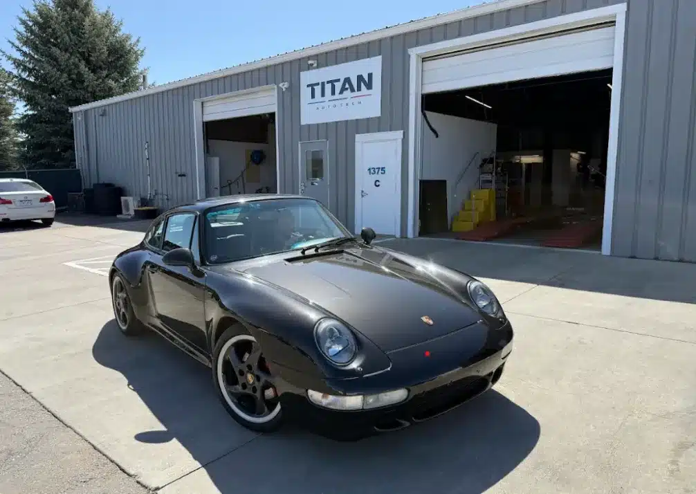Porsche Repair, European Vehicle Repair In Castle Rock CO At Titan Auto Tech European & Import Auto Repair. A black Porsche sports car is parked outside a gray industrial building with TITAN signage above the entrance, and an open garage door reveals a workspace inside.