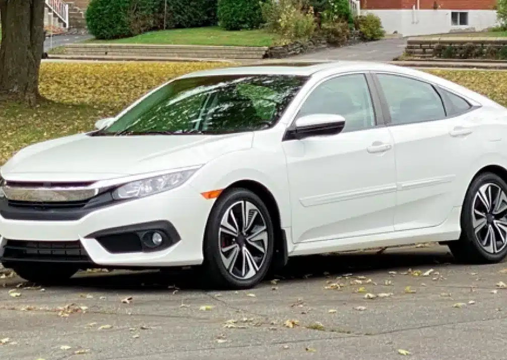 Honda Repair In Castle Rock CO At Titan Auto Tech European & Import Auto Repair. A white sedan car is parked on a paved street with autumn leaves scattered on the ground. There is grass, a large tree, and a brick building with steps in the background.