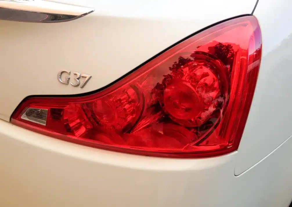 Infiniti Repair In Castle Rock CO At Titan Auto Tech European & Import Auto Repair. Close-up of the rear right taillight and G37 badge on a white car, with reflections of trees visible on the red taillight cover.