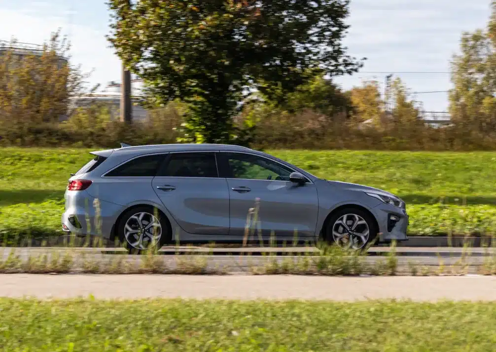 Kia Repair n Castle Rock CO At Titan Auto Tech European & Import Auto Repair. A silver station wagon drives on a road beside green grass and trees, with an industrial structure visible in the background under a partly cloudy sky.