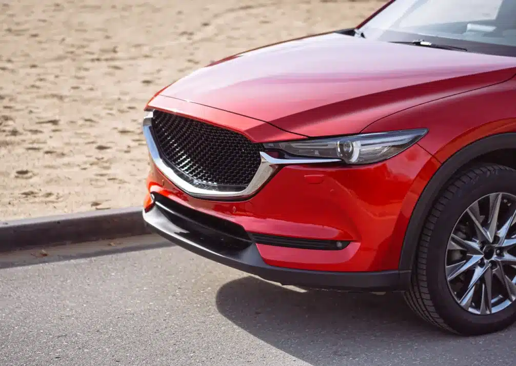 Mazda Repair In Castle Rock CO At Titan Auto Tech European & Import Auto Repair. The front side of a shiny red SUV parked near a sandy area, showing part of the grille, headlight, front bumper, and front wheel.