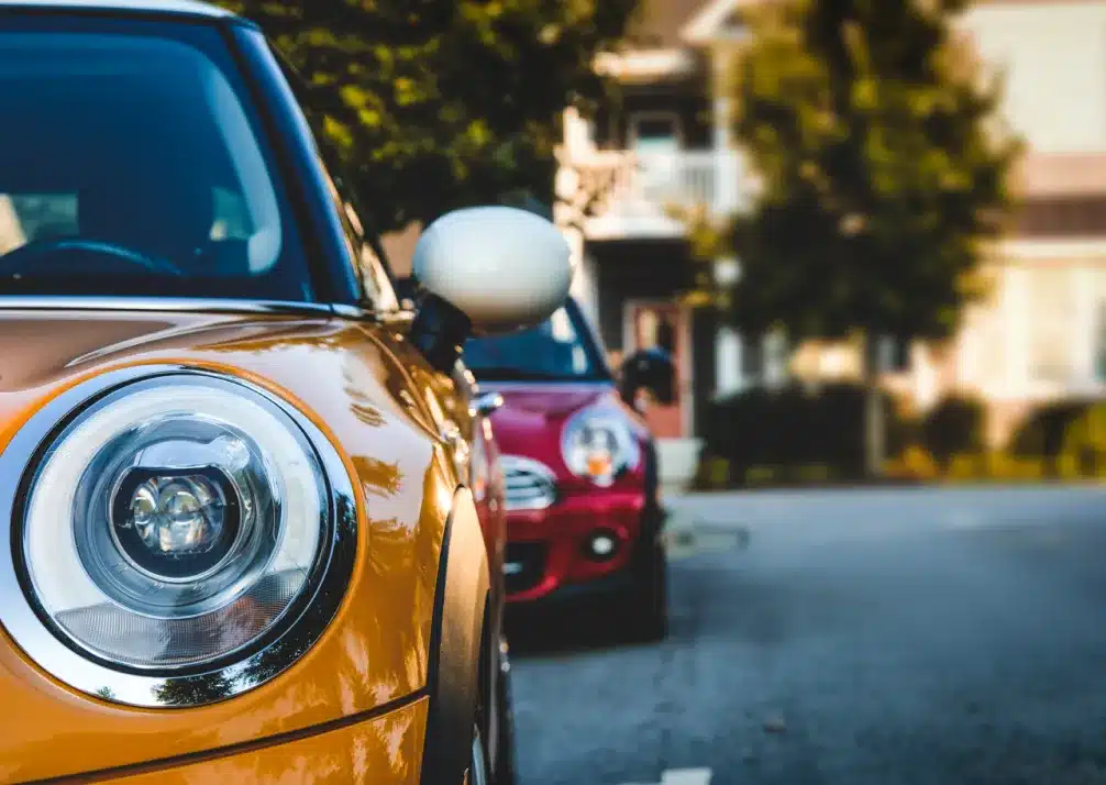 MINI Repair In Castle Rock CO At Titan Auto Tech European & Import Auto Repair. Close-up of the front headlight and side mirror of a yellow car parked on a street, with a red car and suburban houses blurred in the background.