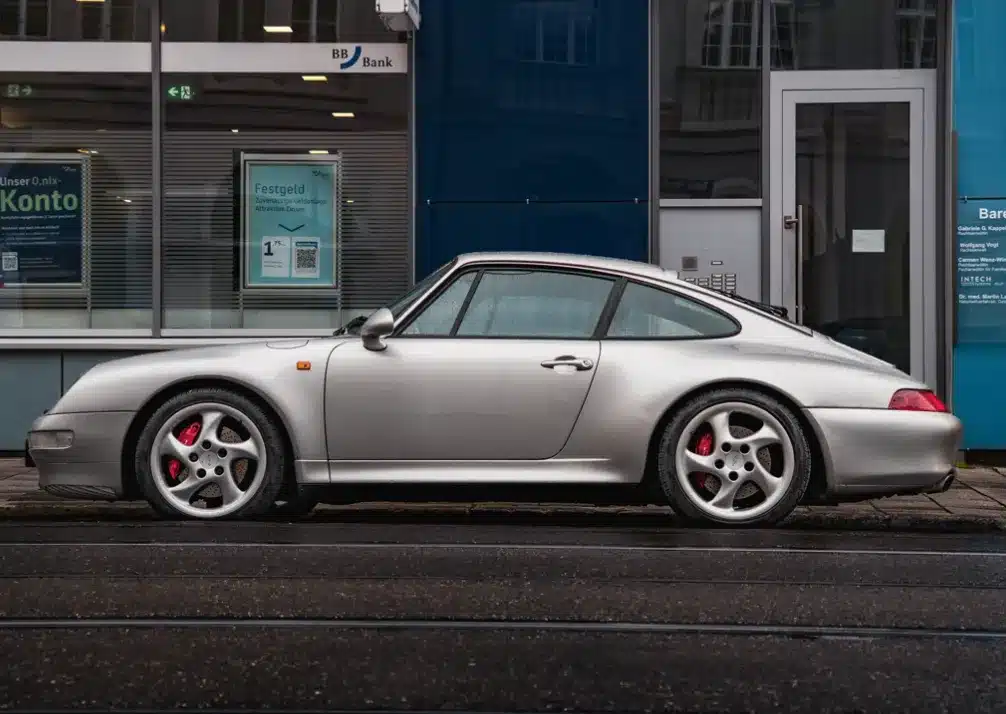 Porsche Repair In Castle Rock CO At Titan Auto Tech European & Import Auto Repair. A silver Porsche 911 sports car is parked on a wet street in front of a glass-walled bank building. The car is viewed from the side, showcasing its sleek design and red brake calipers.