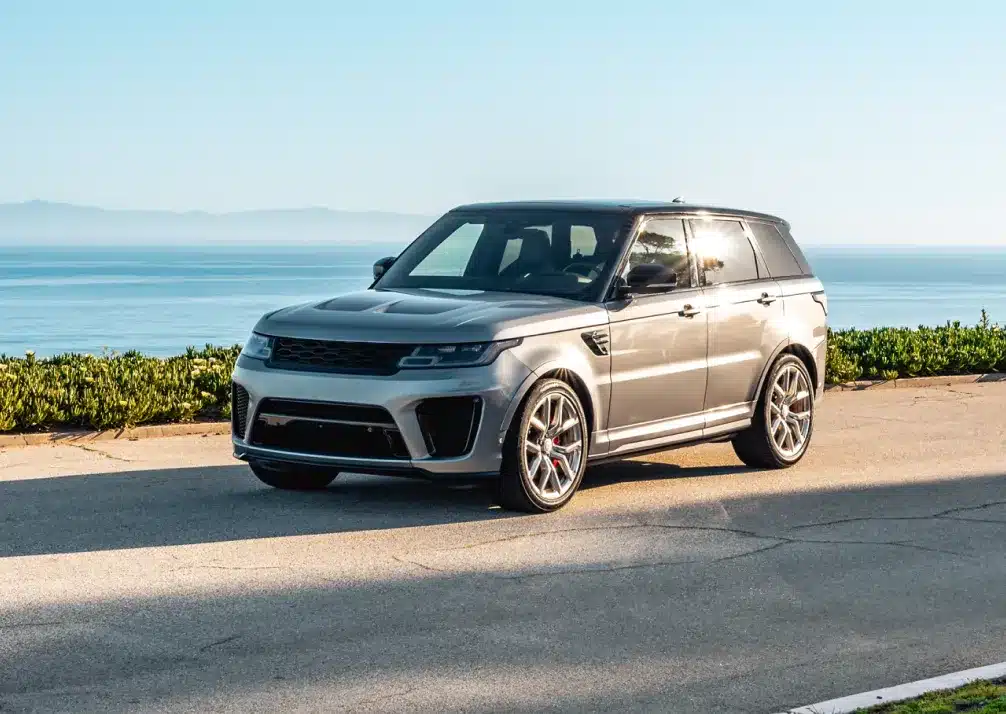 Range Rover Repair In Castle Rock CO At Titan Auto Tech European & Import Auto Repair. A silver SUV is parked on a paved road near the ocean, with clear skies and greenery in the background.