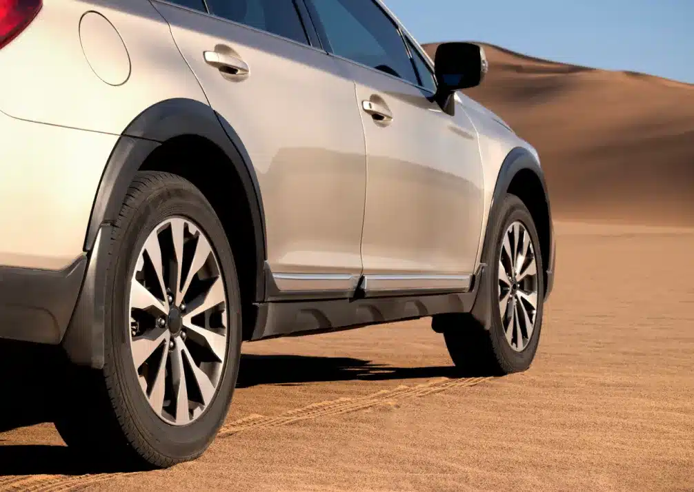 Subaru Repair In Castle Rock CO At Titan Auto Tech European & Import Auto Repair. A beige SUV parked on sandy terrain, with large dunes visible in the background under a clear blue sky. The photo focuses on the rear side and wheels of the vehicle.