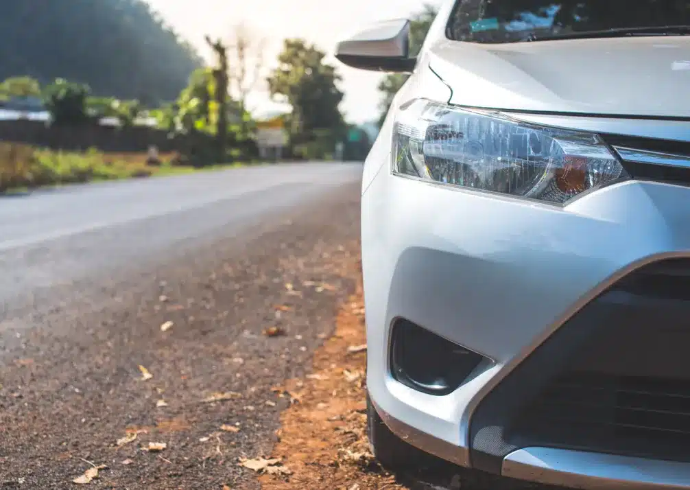 Toyota Repair In Castle Rock CO At Titan Auto Tech European & Import Auto Repair. Close-up of the front right side of a silver car parked beside an empty road, with trees and a hill visible in the background under a bright sky.