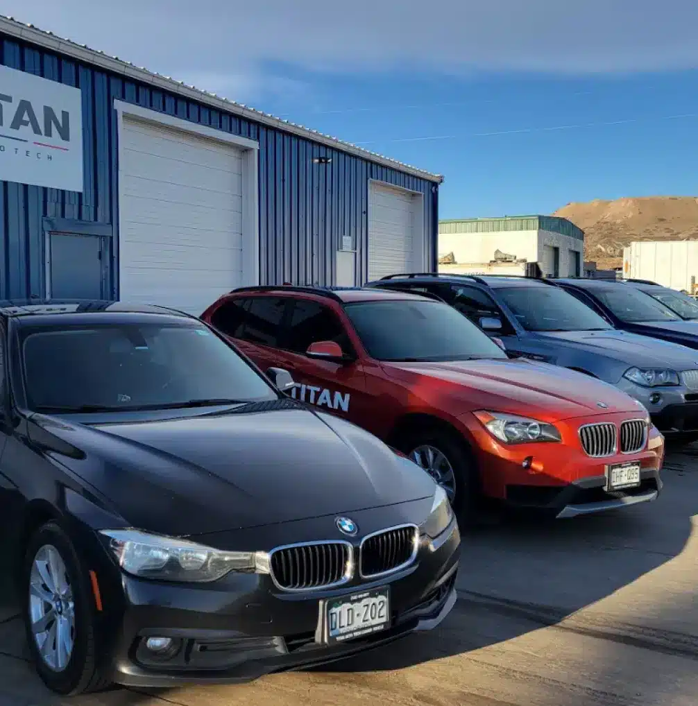 Four BMW vehicles—black, red, gray, and silver—are parked in front of a blue industrial building with white garage doors and a TITAN sign. The sky is mostly clear with some clouds.