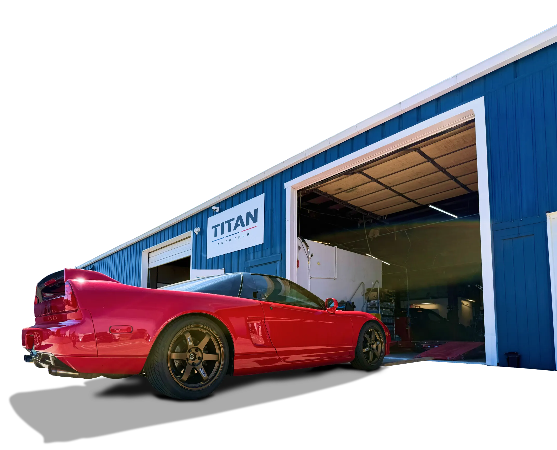 A red sports car with black wheels is parked outside a blue industrial building with a large open garage door and a TITAN sign above it.